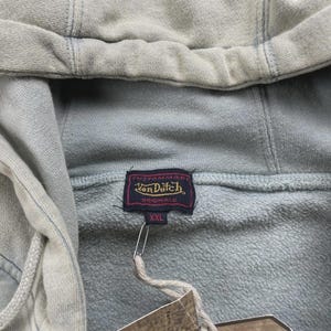 May include: A light green hoodie with a drawstring hood is shown in close-up. The hoodie has a tag with the text "Von Dutch Originals" and the size "XXL". The material looks soft and textured.