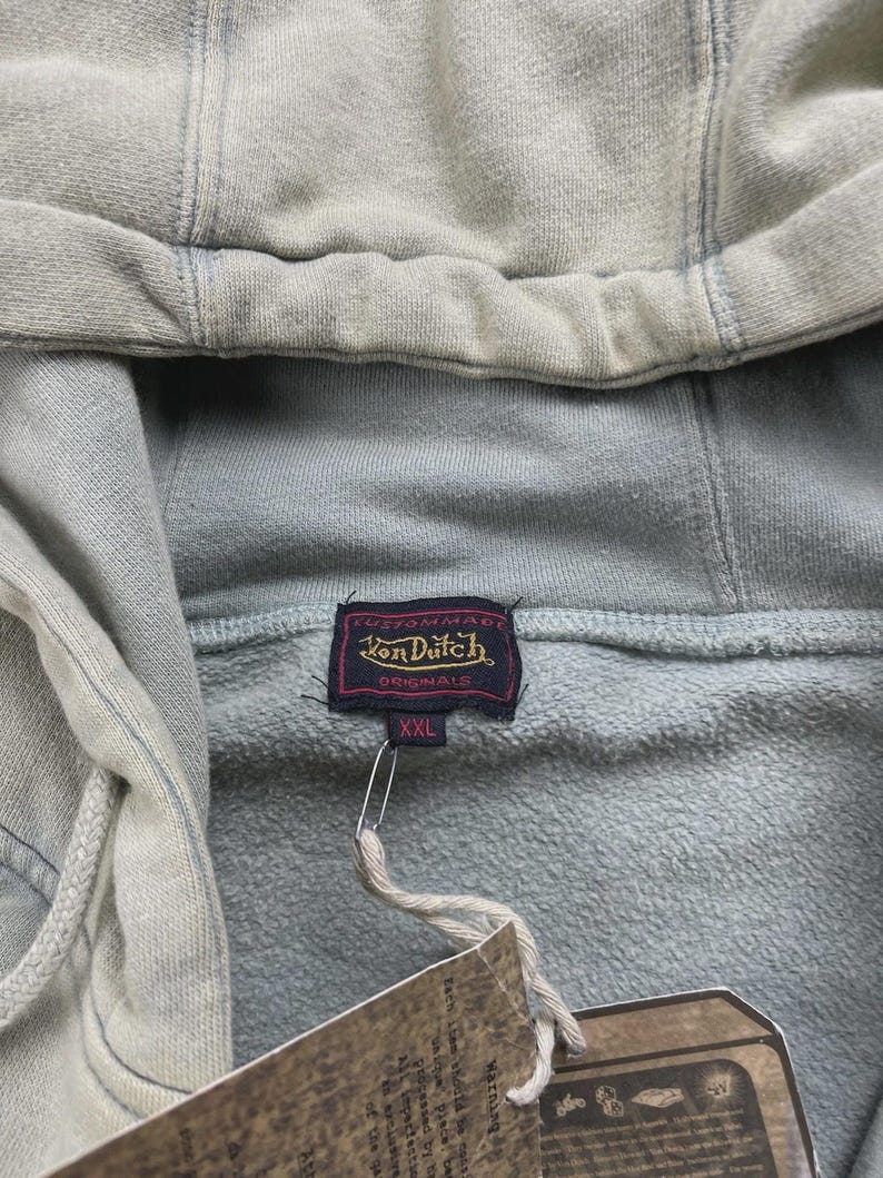 May include: A light green hoodie with a drawstring hood is shown in close-up. The hoodie has a tag with the text "Von Dutch Originals" and the size "XXL". The material looks soft and textured.