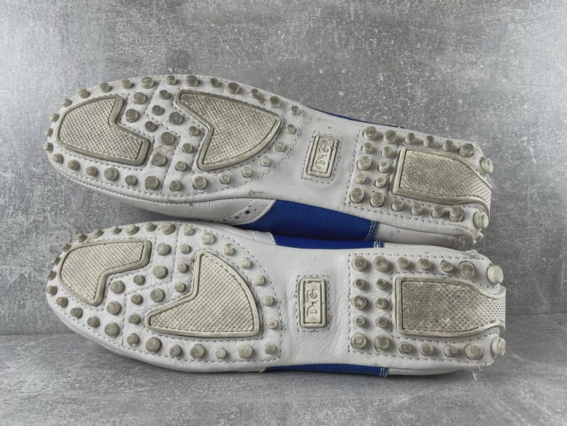 May include: A pair of white and blue leather loafers. The soles are white with small, round, grey rubber studs and textured patches. The blue leather is visible on the sides. The shoes have the "D&G" logo.