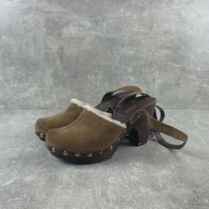 May include: Brown suede clogs with a shearling lining and wooden platform soles. The clogs have a strap around the ankle and are adorned with silver-colored studs. The heels are dark brown and the clogs are set against a gray background.
