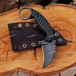 May include: A black karambit knife with a curved blade and a ring at the handle's end, resting on a wooden surface. The knife has a textured grip and is accompanied by a black leather sheath with metal eyelets.