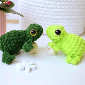 Könnte beinhalten: Zwei Plüschfrosch-Spielzeuge aus weichem, strukturiertem Garn. Ein Frosch ist dunkelwaldgrün mit einem großen gelb-schwarzen Auge, der andere ist hell-limettengrün mit einem großen schwarzen Auge. Jeder Frosch ruht auf einer kleinen geometrischen Holzperle.