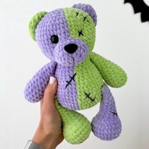 Pode incluir: Um urso de peluche feito de fio macio e texturizado com um design bicolor em roxo claro e verde lima. O urso apresenta olhos de plástico preto, um nariz costurado a preto e detalhes em ponto cruz com fio escuro pelo corpo.