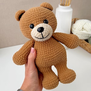 Puede incluir: Un oso de peluche tejido a ganchillo en color marrón con acabado de hilo texturizado, que presenta un hocico color crema, ojos de seguridad de plástico negro y una nariz y boca bordadas en negro. El peluche tiene orejas redondeadas.