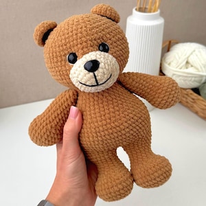 May include: A brown crocheted teddy bear with a textured, plush appearance. The bear features a cream-colored muzzle, two round black plastic eyes, and a small black embroidered nose and mouth. The ears are small, rounded, and match the body.