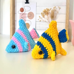 May include: Two handmade crochet fish toys. One is blue with pink stripes, the other is yellow with blue stripes. Both have large black eyes and fins. These soft toys are suitable for children.