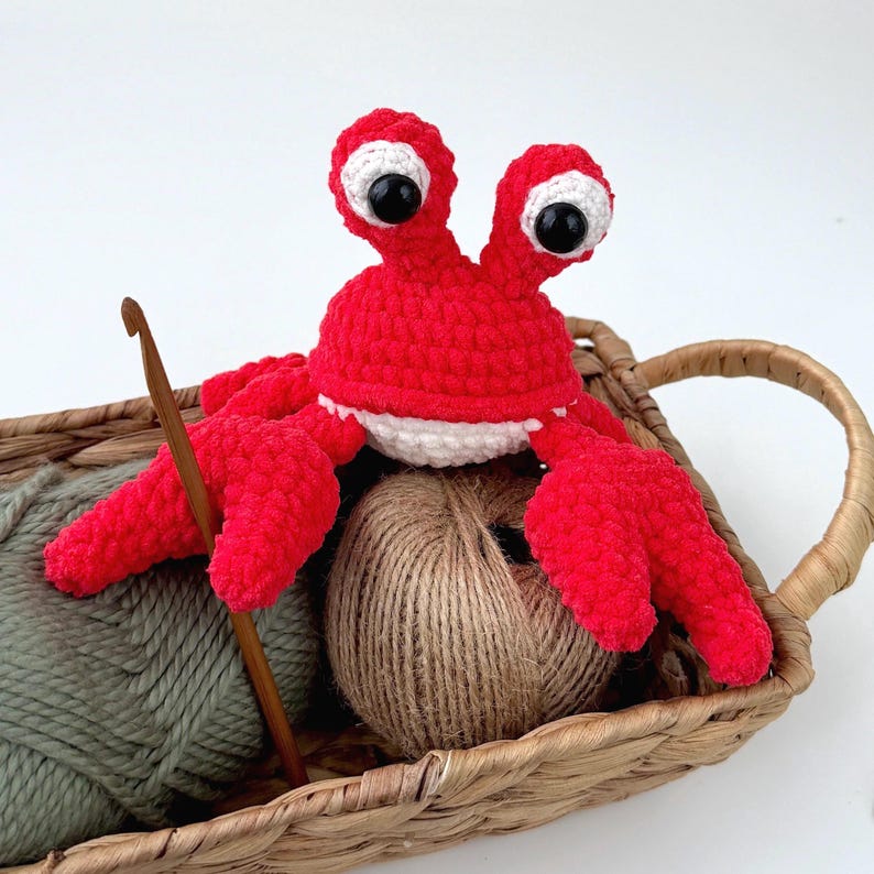 Pode incluir: Um brinquedo de pel&uacute;cia de caranguejo em croch&ecirc; vermelho brilhante com grandes olhos brancos e pretos, posicionado em uma cesta tran&ccedil;ada ao lado de um novelo de barbante marrom, um novelo de l&atilde; verde s&aacute;lvia e uma agulha de croch&ecirc; de madeira.