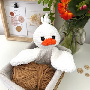 May include: A handmade white crochet duck with an orange beak and black eyes sits in a basket with a ball of brown yarn. The duck has outstretched wings. The basket is lined with gray fabric.