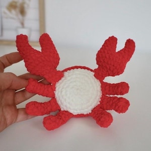 May include: A red crocheted crab toy with white crocheted details. The crab has eight legs and two large claws.