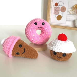 May include: A collection of three crocheted food items: a pink donut with sprinkles, a brown ice cream cone with a pink top, and a brown cupcake with white frosting and a red cherry. Each item has a smiling face.