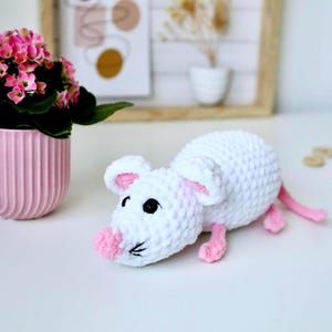May include: A white crocheted mouse with pink ears and nose. The mouse is lying on its belly with its paws outstretched.