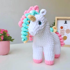 Könnte beinhalten: Ein gehäkeltes Einhorn-Stofftier in Weiß mit strukturierter Garnoberfläche. Es besitzt ein beiges Horn, dunkle Augen mit blauen Akzenten sowie eine Mähne und einen Schweif aus lockigem Garn in Rosa, Mintgrün und Hellgelb. Die Hufe sind hellrosa.
