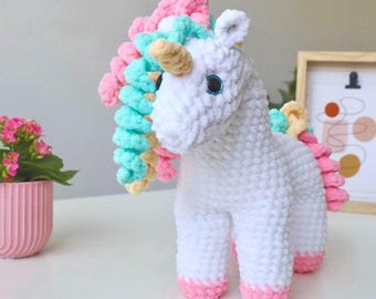 May include: A white crocheted unicorn plush toy with a textured yarn finish. It features a tan horn, dark eyes with blue accents, and a mane and tail made of curly yarn in shades of pink, mint green, and pale yellow. The hooves are light pink.