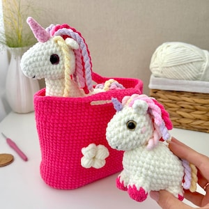 May include: Two white crocheted unicorn plush toys with pink, yellow, and lavender manes and horns. One unicorn is inside a bright pink crocheted basket featuring a white flower applique, while the other is held in front of the basket.
