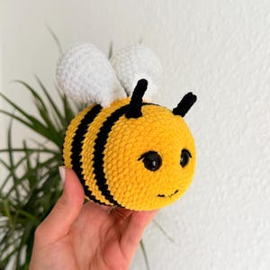 May include: A round, plush crochet bee toy featuring a bright yellow body with thick black horizontal stripes. It has two white textured wings on its back, two small black antennae on top, and two shiny black plastic eyes with a stitched smile.