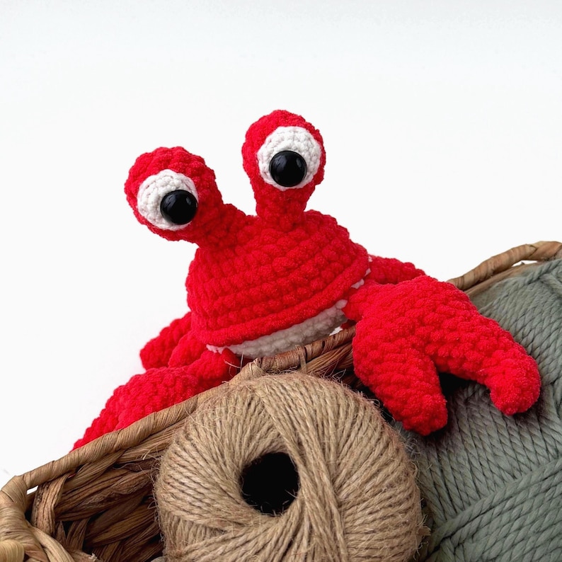 Pode incluir: Um brinquedo de caranguejo de croch&ecirc; vermelho brilhante com dois talos proeminentes encimados por grandes olhos pretos e brancos. O caranguejo est&aacute; posicionado dentro de uma cesta tran&ccedil;ada ao lado de um novelo de barbante e l&atilde; verde.
