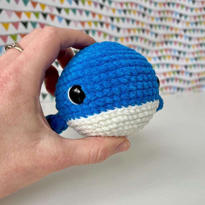 May include: A blue and white crocheted whale plush toy with black eyes. The whale is being held in a hand.