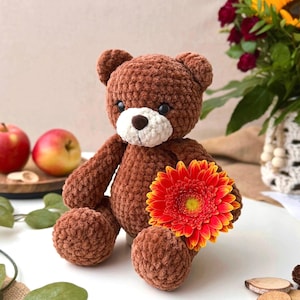 May include: A brown crocheted teddy bear with a white muzzle and a dark brown nose. The bear is holding a vibrant orange and yellow gerbera daisy flower. The plush toy has a soft, textured yarn appearance and round ears, sitting in a seated position.
