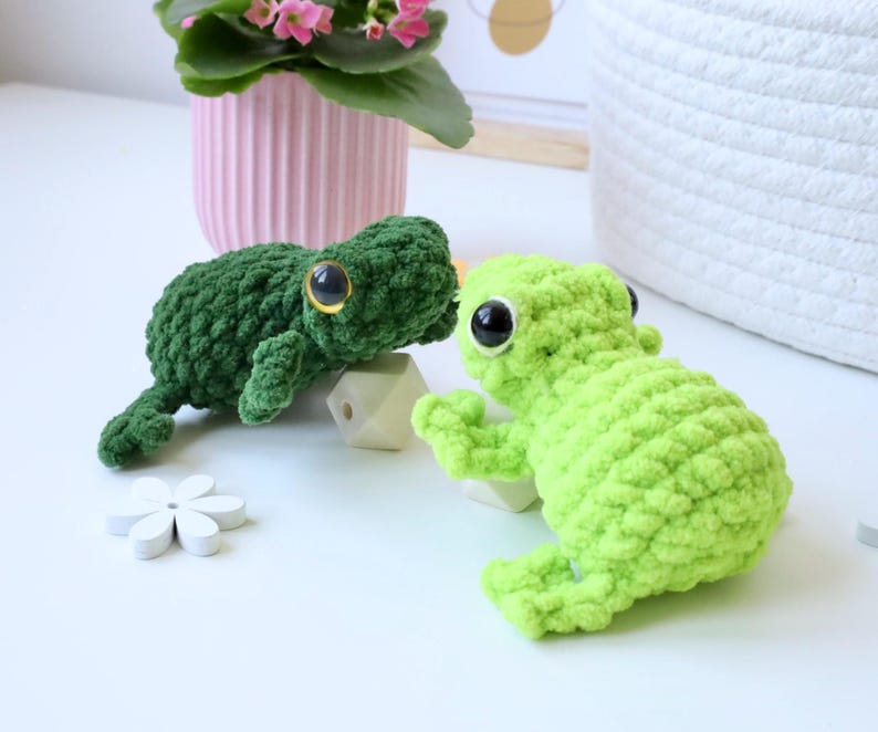 May include: Two crocheted frog plushies, one green and one lime green, with large black eyes. The frogs are sitting on a white surface.