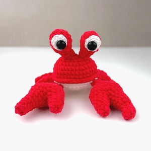 Pode incluir: Um brinquedo de caranguejo feito &agrave; m&atilde;o em croch&ecirc; com fio vermelho brilhante. A pel&uacute;cia apresenta dois talos proeminentes com grandes manchas oculares brancas e olhos de seguran&ccedil;a pretos brilhantes. Tem duas pin&ccedil;as grandes e uma parte inferior branca.