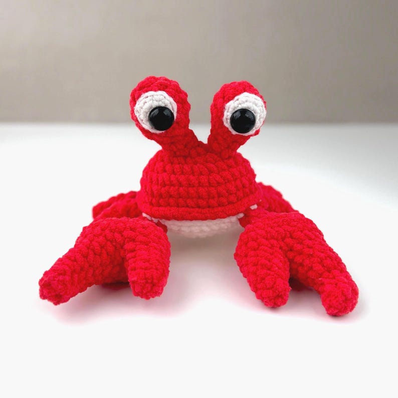 Pode incluir: Um brinquedo de caranguejo feito &agrave; m&atilde;o em croch&ecirc; com fio vermelho brilhante. A pel&uacute;cia apresenta dois talos proeminentes com grandes manchas oculares brancas e olhos de seguran&ccedil;a pretos brilhantes. Tem duas pin&ccedil;as grandes e uma parte inferior branca.