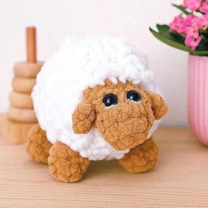 Puede incluir: Un juguete de oveja de ganchillo hecho a mano con un cuerpo blanco y esponjoso y una cara, orejas y patas de color marrón claro. La oveja tiene grandes ojos negros. Un juguete suave y afelpado, ideal para niños.