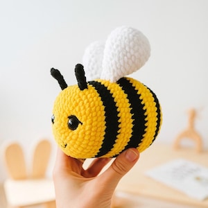 May include: A handmade, plush bee toy. The bee features a yellow body with black stripes, black antennae, and white wings. The toy is held in a person's hand. The bee's eyes are black, and it has a small smile.