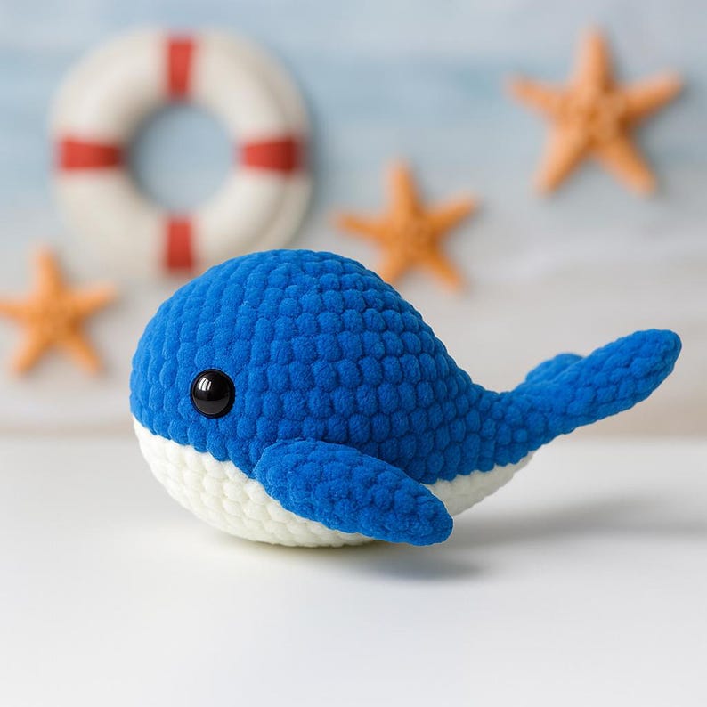 May include: A blue and white crocheted whale plush toy with black eyes. The whale is lying on a white surface.