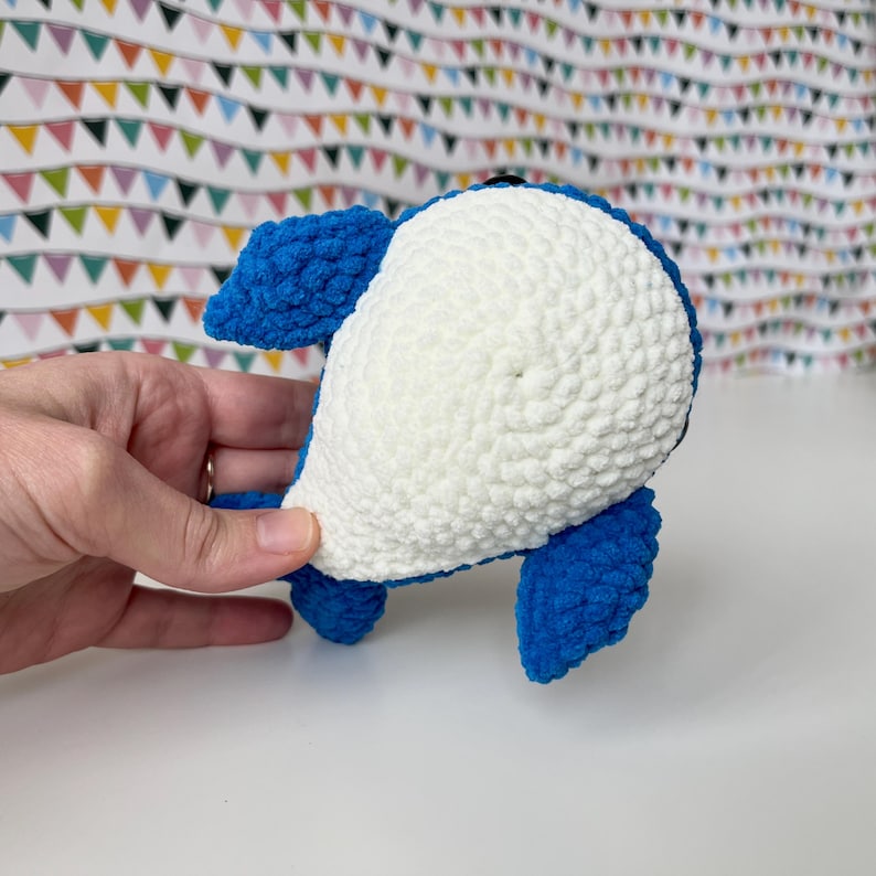May include: A blue and white crocheted whale plush toy. The whale is lying on its back with its tail in the air.