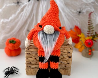 May include: A crocheted gnome featuring a tall orange pointed hat, a round beige nose, and a long grey beard. The gnome has orange and black striped legs and solid black feet, sitting upright with a soft, textured yarn construction.