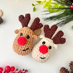 May include: Two handmade crochet reindeer heads. One is tan with brown antlers and a red nose, the other is off-white with brown antlers and a red nose. Both have black eyes and smiling mouths. Festive holiday decor.