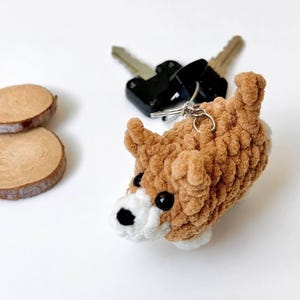 May include: A brown and white crocheted dog keychain made of soft yarn with black plastic eyes and a black nose. The small plush figure is attached to a silver metal keyring, which is connected to a set of black metal keys.