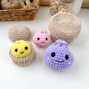 May include: A collection of small, round, crocheted plush toys featuring smiling faces with black bead eyes and pink stitched cheeks. The set includes a yellow dumpling in a beige bowl, a pink dumpling, and a larger purple dumpling.