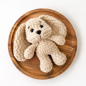 May include: A beige crochet stuffed animal shaped like a dog with long floppy ears. The plush toy features a textured yarn construction, two small black plastic eyes, and a black plastic nose. The dog has a rounded head and a soft body.