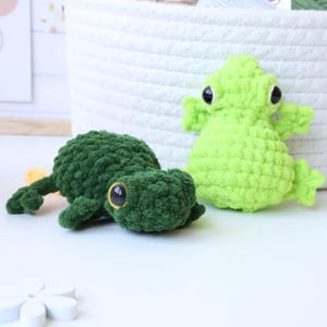 May include: Two crocheted frog plushies, one green and one lime green, with large black eyes. The frogs are sitting on a white surface.