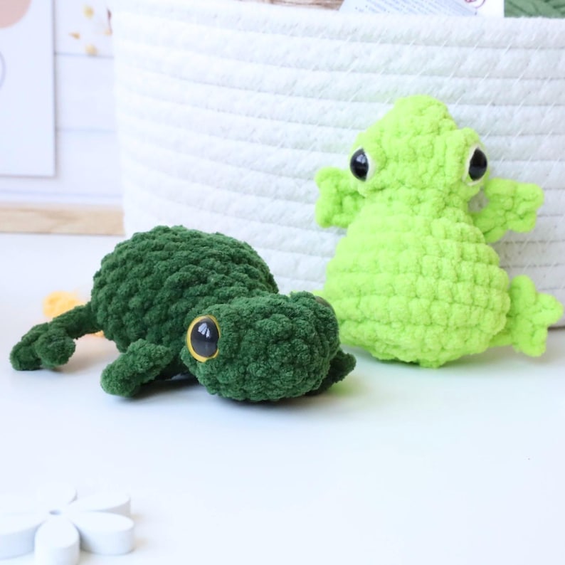 May include: Two crocheted frog plushies, one green and one lime green, with large black eyes. The frogs are sitting on a white surface.