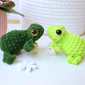 May include: Two crocheted frog plushies, one green and one lime green, with large black eyes. The frogs are sitting on small wooden blocks.