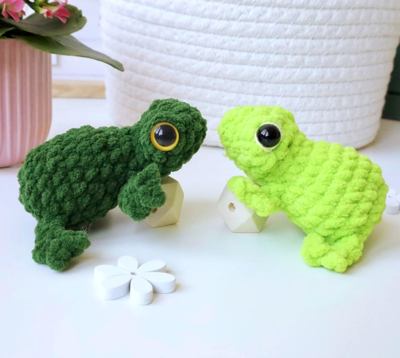 May include: Two crocheted frog plushies, one green and one lime green, with large black eyes. The frogs are sitting on small wooden blocks.