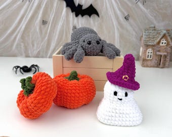 May include: A collection of crocheted items including a gray spider resting on a wooden crate, two orange pumpkins with green stems, and a white ghost wearing a purple witch hat with a small gold-colored buckle detail on the front brim.