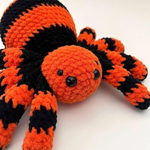 Pode incluir: Uma aranha de pelúcia feita à mão, laranja e preta. A aranha tem um corpo e uma cabeça redondos, com oito pernas. O corpo e as pernas são listrados com laranja e preto. A aranha tem dois olhos pretos. Decoração de Halloween.