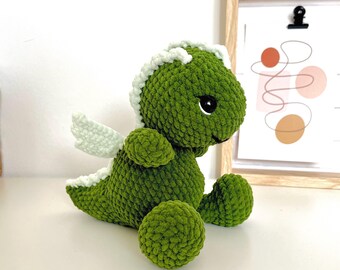 May include: A handmade green crochet dragon with white accents on its head, wings, and spine. The plush toy has large black eyes and a round body. The dragon is sitting on a white surface.