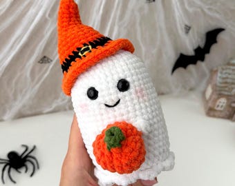 May include: A white crocheted ghost figure featuring a pointed orange witch hat with a black band and a small yellow buckle. The ghost has black bead eyes, a stitched smile, pink cheeks, and is holding a small orange pumpkin with a green stem.