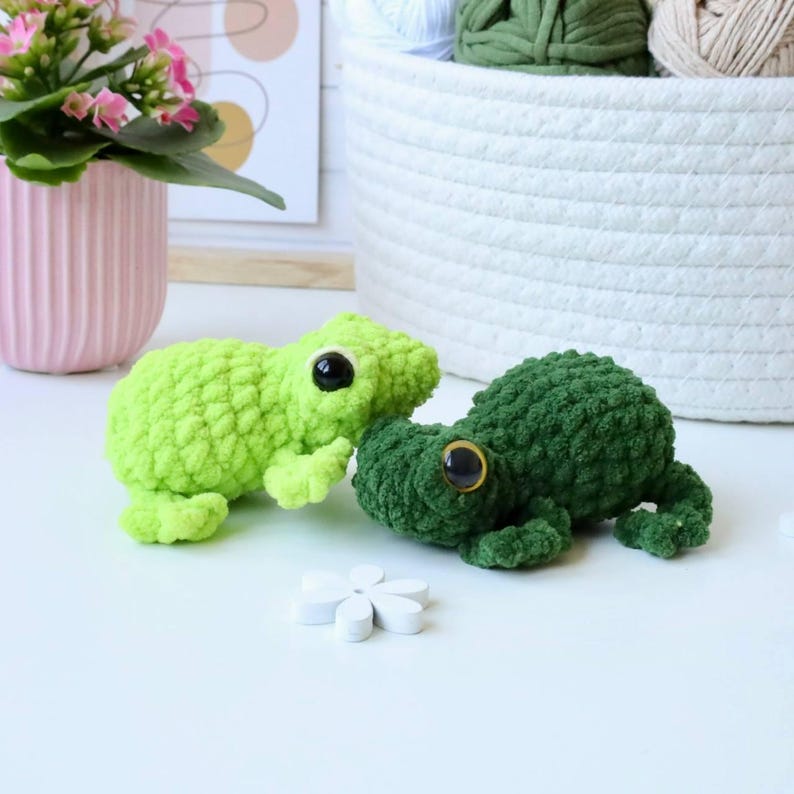 May include: Two crocheted frog plushies, one green and one lime green, with black eyes and a white background.