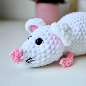 May include: A white crocheted mouse toy with pink accents on the nose, ears, and feet. The mouse has black eyes and a black nose.
