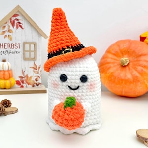 May include: A handmade white crochet ghost wearing an orange witch hat with a black band and buckle. The ghost is holding an orange pumpkin. The ghost has black button eyes and a smiling mouth. A decorative wooden house with fall leaves is in the background.
