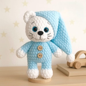 May include: A handmade crochet stuffed animal in the shape of a cat, wearing a light blue onesie and nightcap. The cat has white fur, blue eyes, and a black nose and whiskers. The onesie has two wooden buttons. The toy is standing on a wooden base.