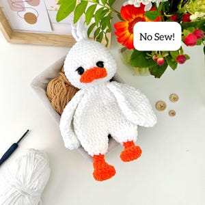May include: A white crocheted duck plush toy with a bright orange beak and feet, black safety eyes, and a tuft of feathers on its head. A white speech bubble overlay contains the black text No Sew! The duck is made of soft, plush yarn.