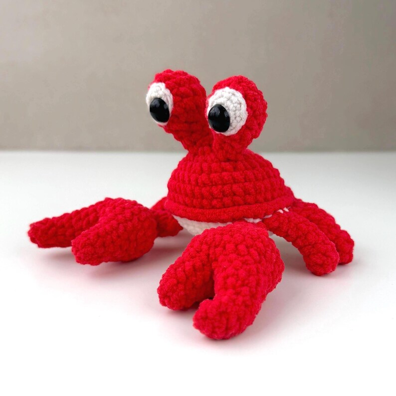 Pode incluir: Um brinquedo de caranguejo em croch&ecirc; vermelho brilhante com um corpo arredondado, duas pin&ccedil;as grandes estendidas para a frente e dois olhos proeminentes em hastes. Os olhos consistem em c&iacute;rculos brancos de croch&ecirc; com olhos de seguran&ccedil;a pretos.