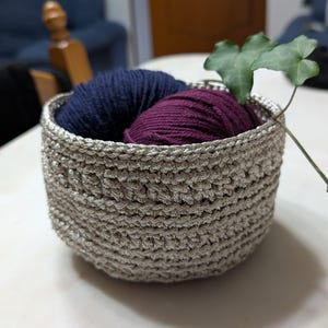 Può includere: Un cesto in crochet argentato con due gomitoli di lana, uno blu scuro e uno bordeaux, all'interno. Una foglia verde sporge dal cesto.