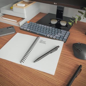 a well-organized workspace with a computer keyboard, a notebook, a pen, a mouse, and a pair of glasses on a wooden desk.